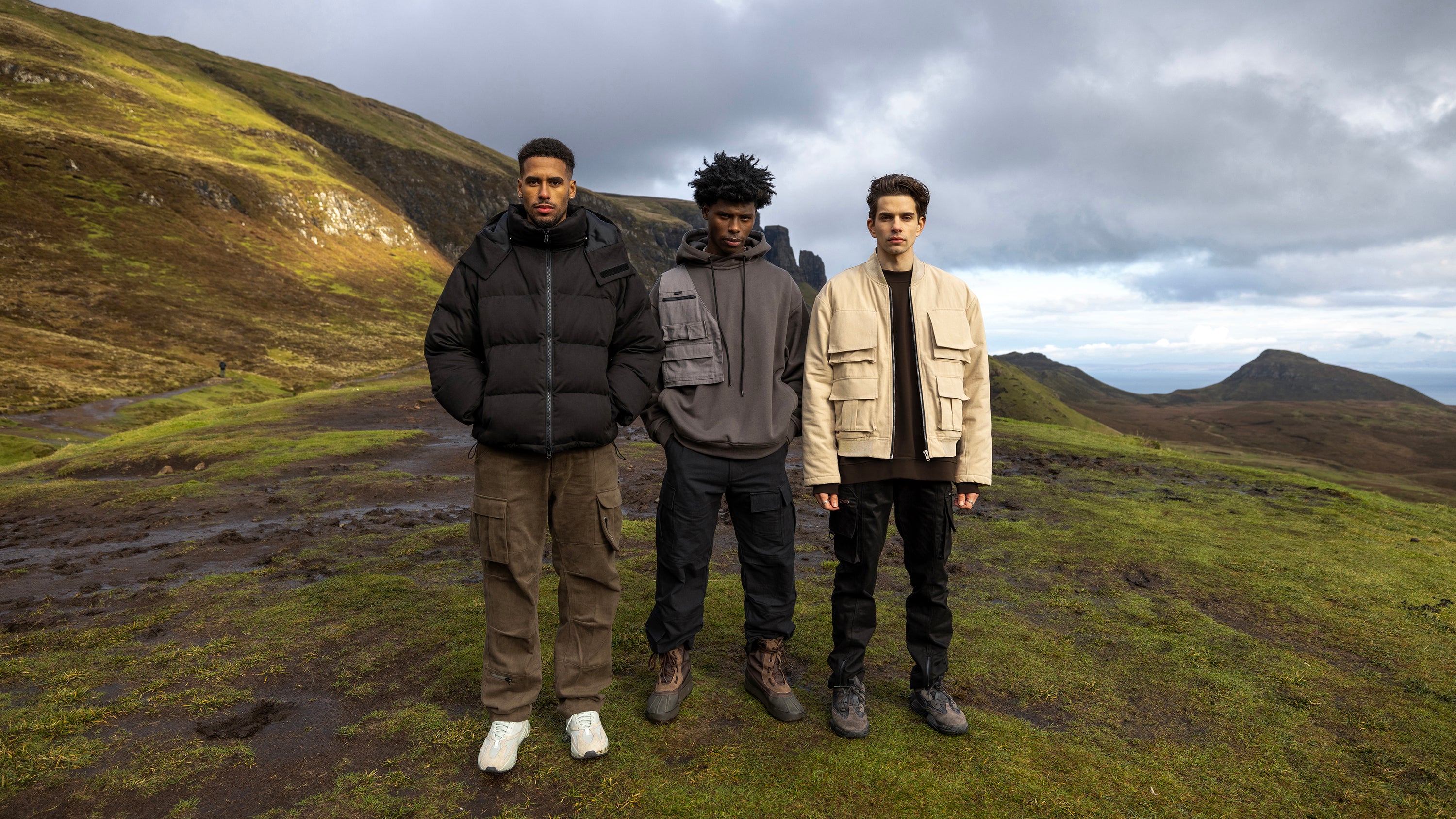 Three male models in black cargo pants and jackets stand atop the Isle of Skye.