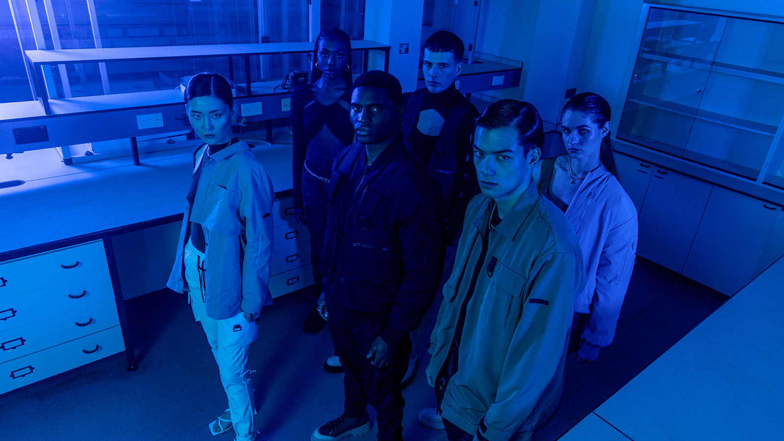 Six models wearing cargo pants and jackets pose in a laboratory.